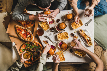 Lockdown fast food dinner from delivery service. Flat-lay of friends sitting and having beer quarantine party with burgers, french fries, sandwiches, pizza and salad over table background, top view - 413171133
