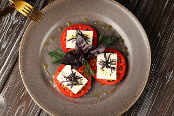 An overhead view of a cold appetizer of ripe red tomatoes, feta cheese and purple basil, seasoned with olive oil and black pepper.