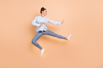 Fototapeta na wymiar Full size profile photo of optimistic nice brunette lady jump karate wear blue sweater jeans sneakers isolated on beige background