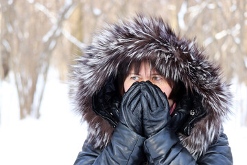 Frozen woman in leather coat with fur hood standing outdoors with her gloved hands covering her...