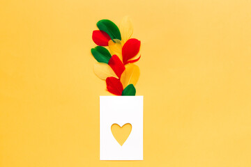 Blank white card in shape of heart, on yellow background, with colored feathers, top view. Greeting card template. Love concept. Copy space.