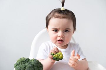 Baby girl sitting in baby chair eating broccoli on white background.