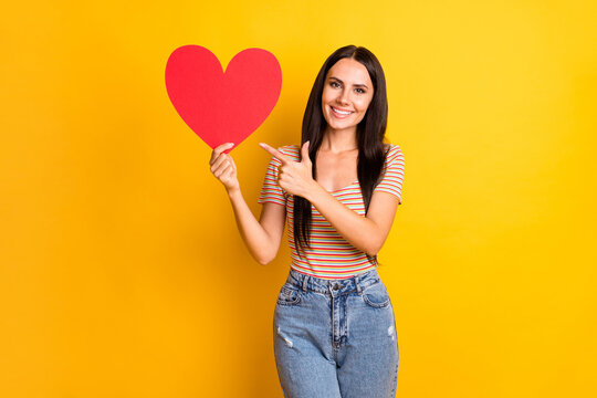 Photo portrait of curious girl pointing finger heart shaped red card wearing casual outfit isolated on vibrant yellow color background