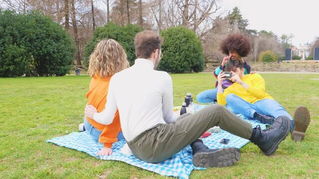 Four cheerful friends diverse multiethnic having fun doing pic nic in a park outdoors using smartphone taking photos making memories