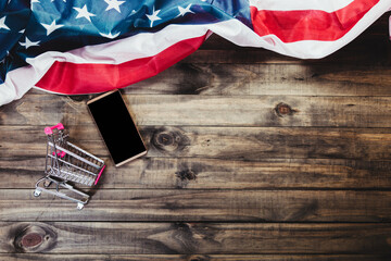 Online shopping concept - Image of a smarthphone or cell phone and a shopping cart on a wooden background with a US flag.