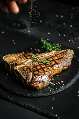 Gourmet t-bone steak or aged wagyu porterhouse grilled beef steak in American meat restaurant. Chef sprinkles with seasoning in a freeze motion on black background. vertical image