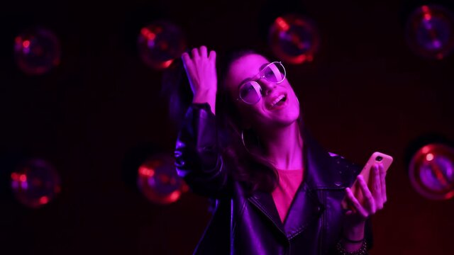 Beautiful brunette girl in club atmosphere. Happy female in sunglasses dancing in room in the pink and blue neon light, looking on cellphone and smiling. Close up. Slow motion. stylish outfit.