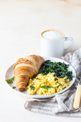 Croissant with scrambled eggs and spinach on plate served with coffee, light concrete background. Breakfast.