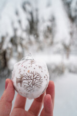 vertical view of christmas ornament with a reindeer hold by a woman hand. Background with copyspace of a pine covered with snow outdoors. Seasonal winter december concept.
