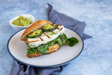Croissant sandwich with grilled halloumi cheese, arugula and grilled zucchini, blue concrete background. Healthy breakfast or lunch.