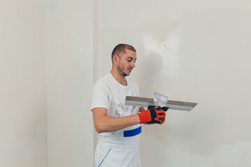 A man worker plasters the wall with a spatula and applies a cement mix solution. The repairman lays the plaster on the drywall.