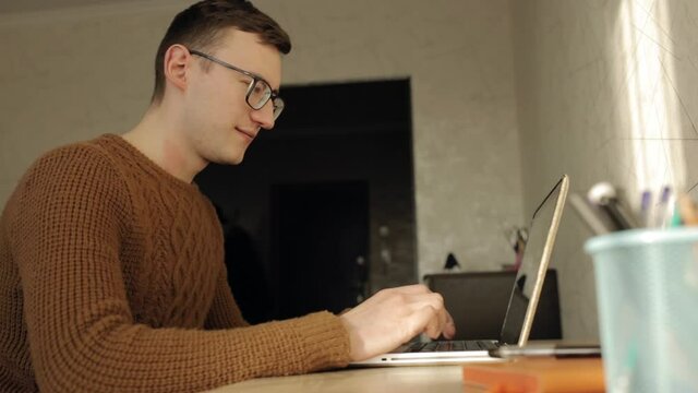 Side view of exhausted male entrepreneur in glasses rubbing eyes while sitting at table and typing on laptop during distant work from home office 
