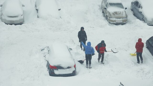 People in a backpack clearing their cars of snow with shovels. A group of people clean the parking lot near their house from the snow. Heavy snowfall. Snow drifts.