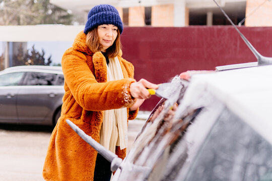 Winter portrait of a mature woman in red fur coat faux fur cleaning snow from rear glass of a car. Beauty red haired senior female smiling s and cheerfully cleans the snow. Elderly woman outdoors.