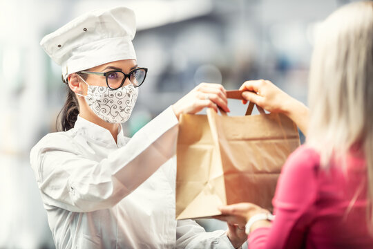 Chef wearing hat, glasses and face mask hands a paper bag with delivery food to a customer