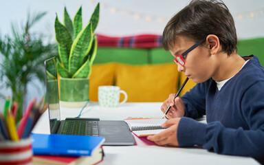 Boy having online classes at home writing in a notebook and using laptop