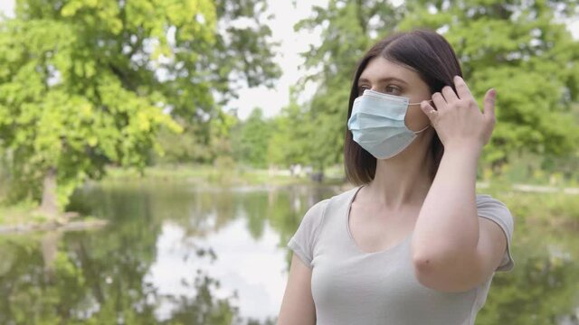 A young Caucasian woman in a face mask relaxes in a park - a pond and trees in the blurry background