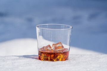 Glass of whiskey with ice on a bed of snow and white background, close up