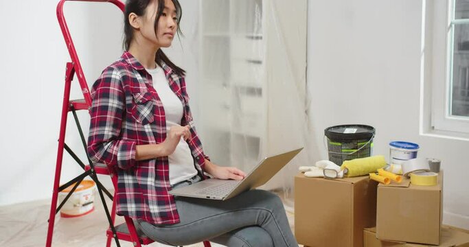 Portrait of concentrated Asian young beautiful woman sitting on red ladder in room with boxes during renovation tapping on laptop browsing online choosing new decor for house, designing home concept