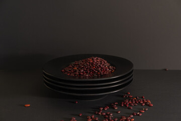 Still life of a stack of black plates with red beans on a black background.