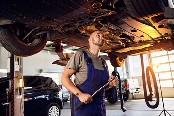 Fototapeta na wymiar Handsome auto mechanic checking running gear of automobile on service station. Cheerful male worker fixing problem with car. Concept of problem solving