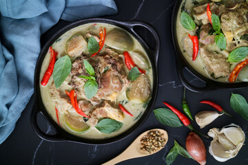 Top view of traditional Thai green curry with lamb and eggplant with its ingredients and spices such as grain de coriander, garlic, shallot, red chili and fresh sweet basilic leaves in a black bowl 