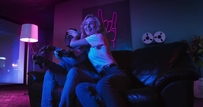 Young man and woman are playing video games together using controllers. They are sitting on the sofa in the living room with red and blue neon lighting. Girl wins and celebrates victory.