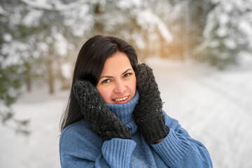 A young smiling brunette woman in a blue sweater and black mittens holds her head with hands in a snowy winter forest. Selective focus.