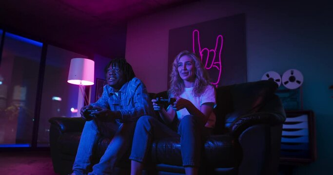 Young man and woman are playing video games together using controllers. They are sitting on the sofa in the living room with red and blue neon lighting. Girl wins and celebrates victory. Dynamic shot.