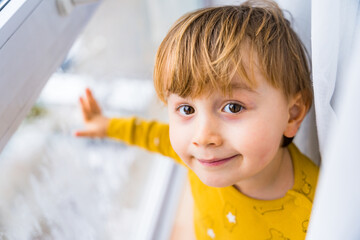 Portrait of a smiling little boy staying near the window and looking at the camera. Toddler staying at home in rainy weather.