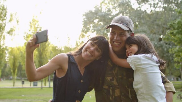 Happy military couple of parents and cute child taking selfie on smartphone in city park. Soldier dad holding kid in arms and smiling at camera. Family reunion or returning home concept