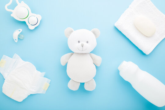 White baby diaper, towel, soap, bottle of talcum powder, soother and rattle toy around little teddy bear on light blue table background. Pastel color. Closeup. Top down view.