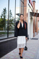 Fototapeta premium Young brunette girl with red pony tail, wearing stylish black jacket and white silk dress, standing in front of modern glass building. Pretty business woman on lunch break. Female portrait in city.