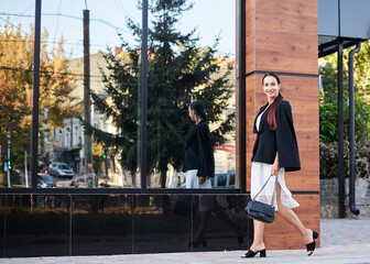 Young brunette girl with red pony tail, wearing stylish black jacket and white silk dress, walking by modern glass building. Pretty business woman on lunch break. Female portrait in city.