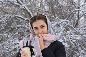 Portrait of  young woman with beautiful blue eyes. Lady on walk in  winter snowy forest drinks hot tea from mug and holds scarf with  hand