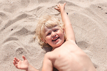 Little boy lying on the sand and playing on the beach of Mediterranean sea. Summertime, vacation, travel concept.