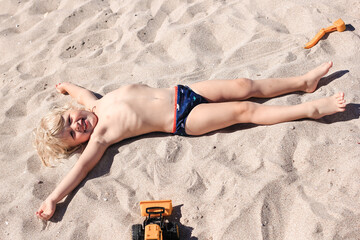 Little boy lying on the sand and playing on the beach of Mediterranean sea. Summertime, vacation,...