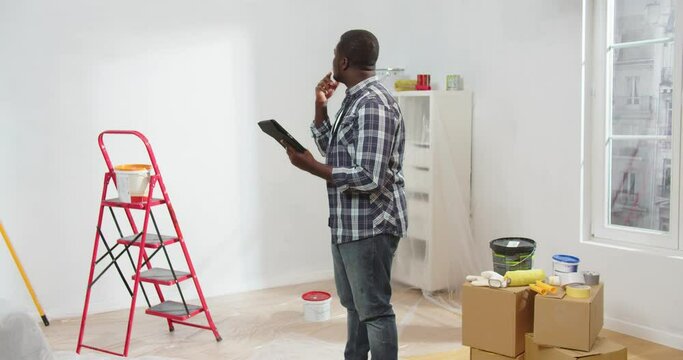 Rear of African American man typing on tablet in room during home renovation, young busy guy repairman browsing online on device choosing new design and decor for apartment, design concept
