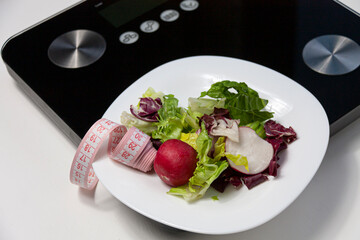 some salad and a pink centimeter on white plate standing on black scales on white background