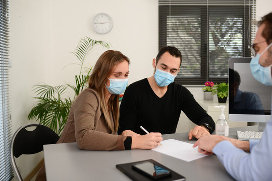 Young Couple Signing Documents In A Business Bank Insurance Office Wearing  Protective Surgical Mask With A Professional Agent During Covid 19 Pandemic