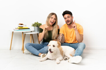Young caucasian couple with dog staying at home thinking an idea pointing the finger up