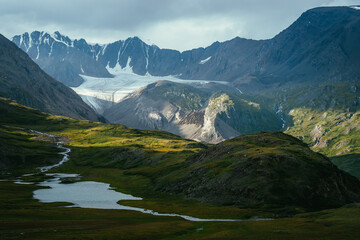 Atmospheric alpine landscape with mountain lake in green valley and glacier under cloudy sky. Awesome highland scenery with beautiful glacial lake among sunlit hills and rocks against mountain range.