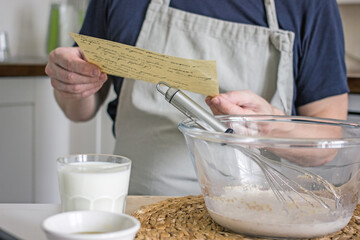 A man without a face in an apron kneads dough for homemade bread according to a recipe written on a piece of paper, which he holds in his hands. Cooking at home learning concept, authentic hobby