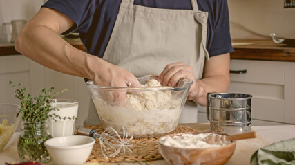 A faceless man in an apron in his kitchen at the kitchen table kneads bread dough in a glass bowl with his hands. Home authentic hobby, home baker. Baking bread with your own hands