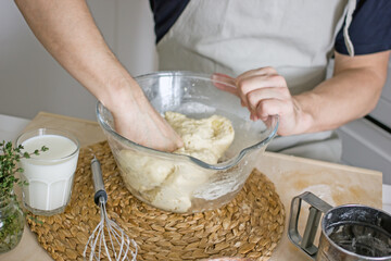 A faceless man in an apron in his kitchen at the kitchen table kneads bread dough in a glass bowl with his hands. Home authentic hobby, home baker. Baking bread with your own hands