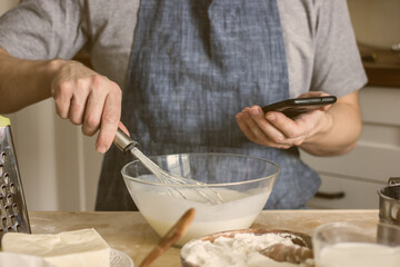 A  baker with no face in an apron makes homemade bread on the kitchen table and looks at his phone for clues. Authentic hobby home baker. The concept of cooking with an electronic assistant