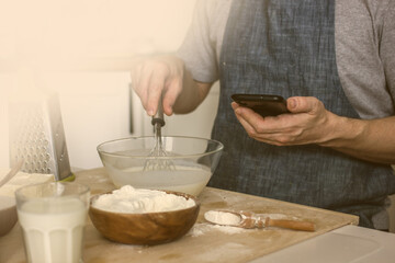 A  baker with no face in an apron makes homemade bread on the kitchen table and looks at his phone for clues. Authentic hobby home baker. The concept of cooking with an electronic assistant