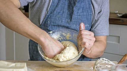 A faceless man in an apron in his kitchen at the kitchen table kneads bread dough in a glass bowl with his hands. Home authentic hobby, home baker. Baking bread with your own hands