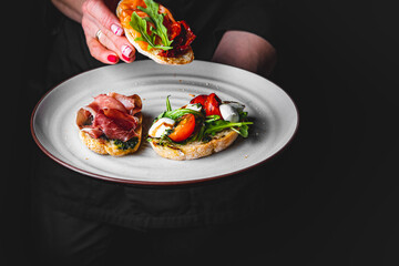 chef in a black suit holds in his hands plate with Bruschetta on Dark grey black background