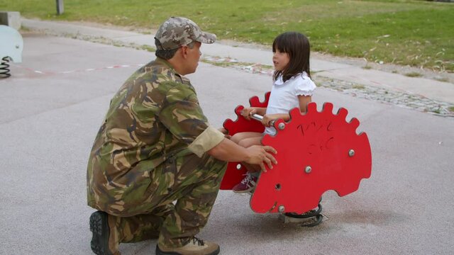 Girl enjoying time with military daddy, riding spring rocking hedgehog on playground. Male soldier playing with active little daughter outdoors on vacation. Parenthood or childhood concept
